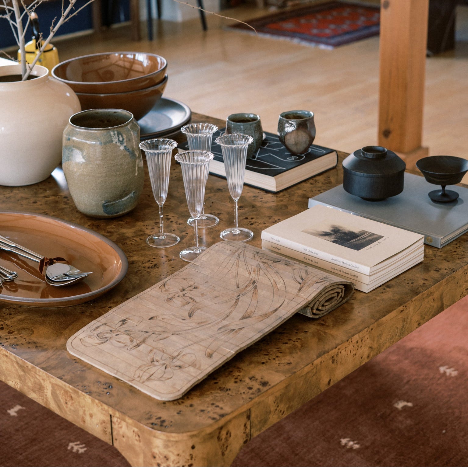 Table setting with ceramic items, glasses, and books in a room with wooden floor and furniture.